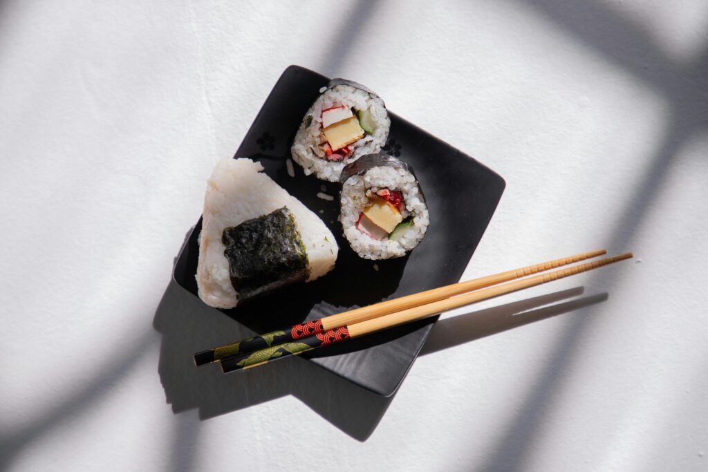 Overhead shot of sushi rolls and onigiri on a black plate with chopsticks.
